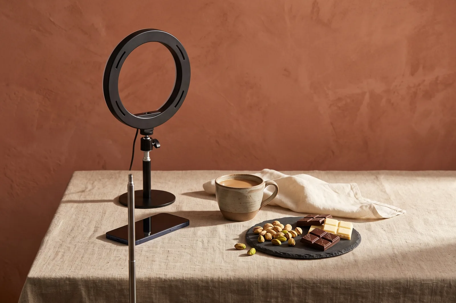 A home content studio corner with a ring light, a phone, coffee and squares of chocolate on a linen table